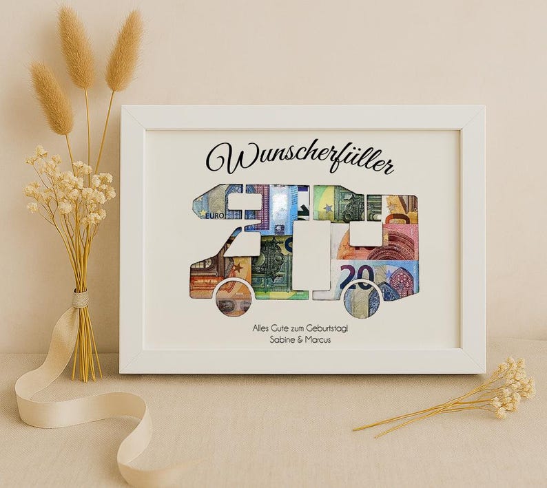 May include: White framed picture with a camper van cutout filled with euro bills. The text "Wunschersch&uuml;ller" is at the top of the frame and "Alles Gute zum Geburtstag Sabine & Marcus" is at the bottom.