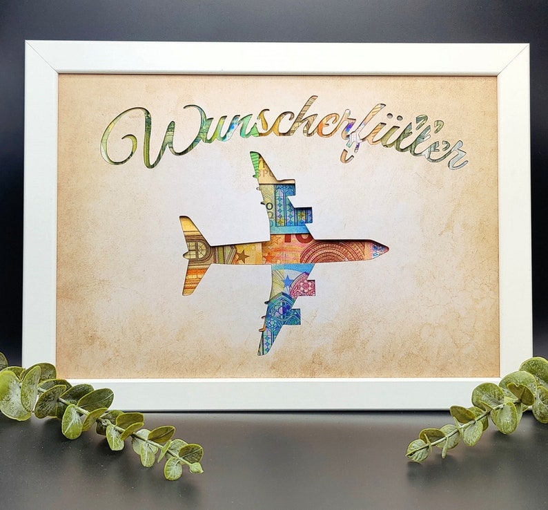 May include: A white frame with a cutout of an airplane made of euro bills. The text "Wunsch erf&uuml;llt" is written above the airplane.