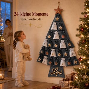 Peut inclure: Un calendrier de l'Avent mural en forme de sapin de Noël en feutrine gris foncé, avec 24 petites pochettes numérotées blanches et gris foncé. Une étoile cuivrée orne le sommet et le prénom Leni est inscrit en lettres cursives à la base.