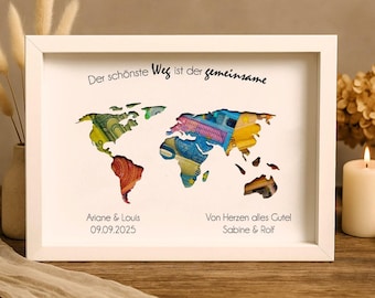 May include: A white picture frame with a world map cutout filled with colorful paper money. The text "Der schönste Weg ist der gemeinsame" is at the top of the frame. The names "Ariane & Louis" and "09.09.2025" are at the bottom left of the frame. The text "Von Herzen alles Gute" and "Sabine & Rolf" are at the bottom right of the frame.
