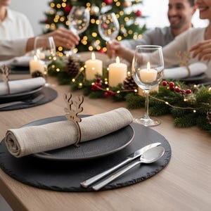 May include: A festive table setting with black plates, linen napkins, and silver cutlery. Wooden reindeer napkin rings and lit candles add to the holiday ambiance. A Christmas tree with lights is in the background.