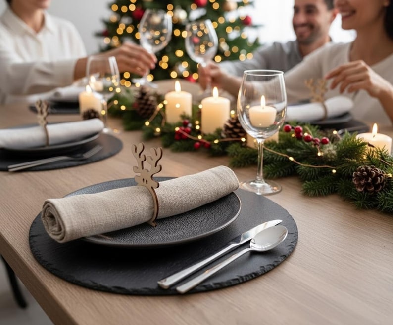May include: A festive table setting with black plates, linen napkins, and silver cutlery. Wooden reindeer napkin rings and lit candles add to the holiday ambiance. A Christmas tree with lights is in the background.