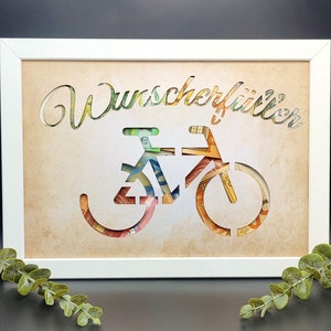 May include: A white frame with a brown background and a cutout of a bicycle made from money. The text "Wunscherrittler" is written above the bicycle.