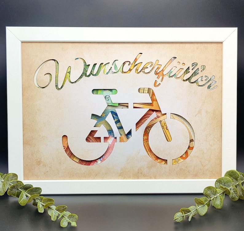 May include: A white frame with a brown background and a cutout of a bicycle made from money. The text "Wunscherrittler" is written above the bicycle.