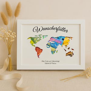 May include: White framed picture with a world map cutout filled with colorful paper money. The text "Wunschersch&uuml;ller" is at the top of the frame and "Alles Gute zum Geburtstag Sabine & Marcus" is at the bottom.