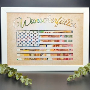 May include: A white framed picture with a brown background. The picture features a cutout of an American flag with the word "Wunsch erf&uuml;llt" written above it. The flag is made of euro bills.