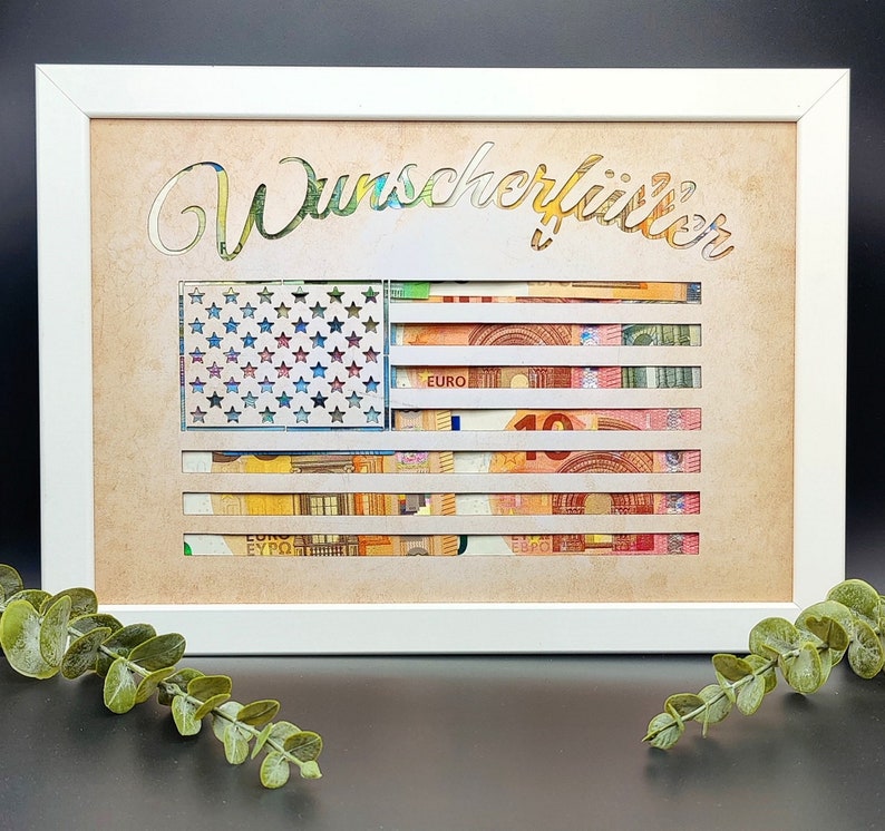 May include: A white framed picture with a brown background. The picture features a cutout of an American flag with the word "Wunsch erf&uuml;llt" written above it. The flag is made of euro bills.