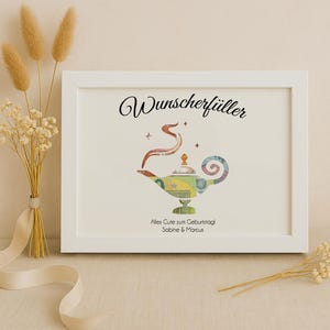 May include: A white frame with a white mat and a picture of a genie lamp made of money. The text "Wunschersch&uuml;ller" is at the top of the picture. The text "Alles Gute zum Geburtstag Sabine & Marcus" is at the bottom of the picture.