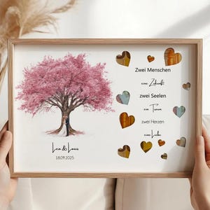 May include: A framed print featuring a pink flowering tree with a couple standing beneath it. The print includes the names "Lea & Louis" and the date "18.09.2025". Decorative hearts and German text are also present.
