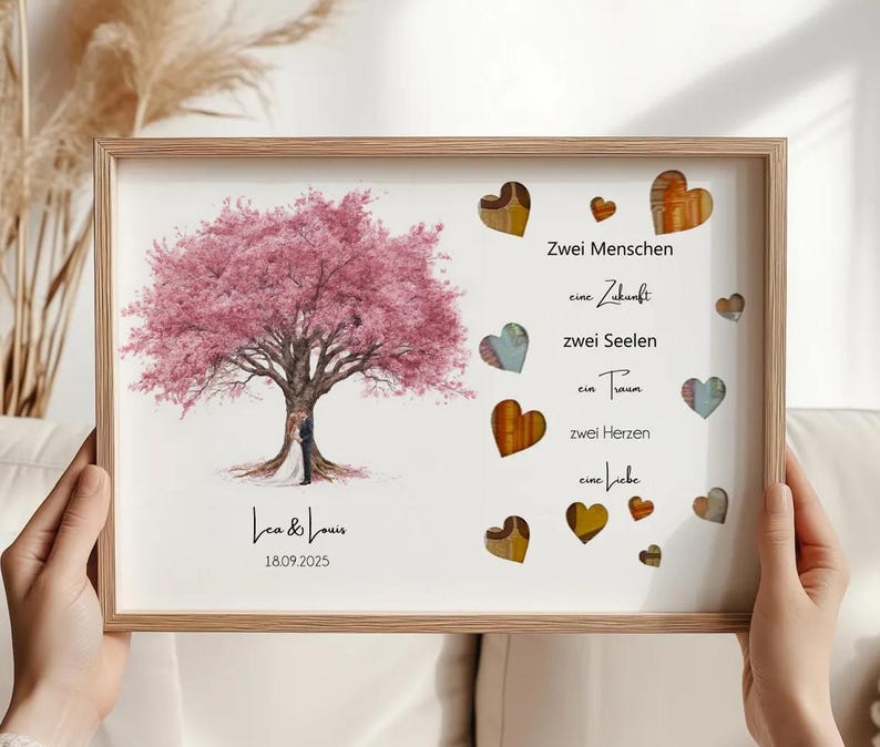 May include: A framed print featuring a pink flowering tree with a couple standing beneath it. The print includes the names "Lea & Louis" and the date "18.09.2025". Decorative hearts and German text are also present.