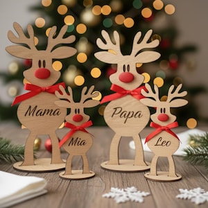 Puede incluir: Decoraciones navideñas de familia de renos de madera. Cada reno tiene una nariz roja y una cinta roja. Los renos están grabados con las palabras "Mama", "Papa", "Mia" y "Leo".