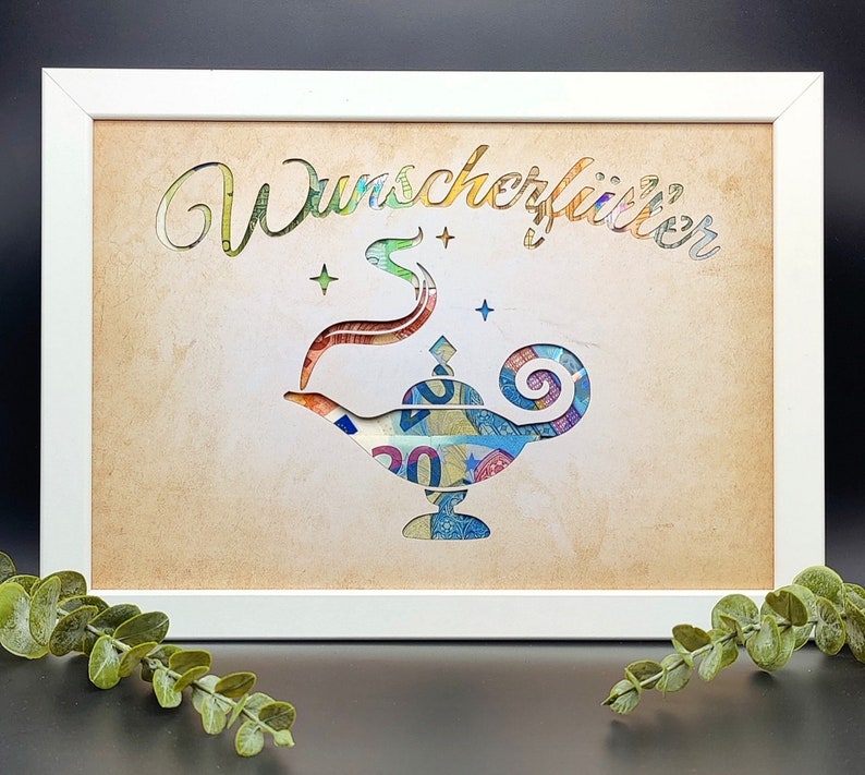 May include: A white frame with a tan background and a cutout of a genie lamp made from money. The text "Wunsch erf&uuml;llt" is written in a decorative font above the lamp.