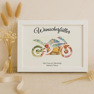 May include: A white frame with a white mat and a motorcycle silhouette made of money inside. The text "Wunschersch&uuml;ller" is at the top of the frame. The text "Alles Gute zum Geburtstag Sabine & Marcus" is at the bottom of the frame.