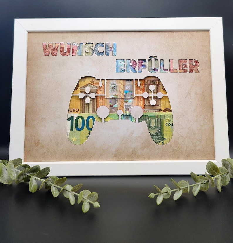 May include: A white rectangular frame containing a tan mat with the cutout text WUNSCH ERF&Uuml;LLER and a game controller shape. Behind the cutouts, Euro banknotes are visible, showing various colors and the number 100 on a green bill.