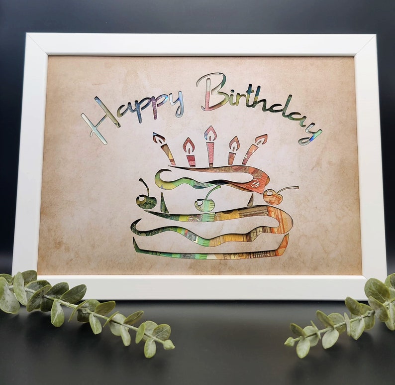 Puede incluir: Una imagen enmarcada en blanco con un fondo marr&oacute;n. La imagen tiene un recorte de un pastel de cumplea&ntilde;os hecho de dinero con las palabras "Happy Birthday" encima.