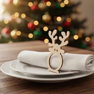 May include: A wooden reindeer napkin ring sits atop a white rolled napkin, placed on a white plate. The table setting is on a wooden surface, with a blurred Christmas tree in the background.