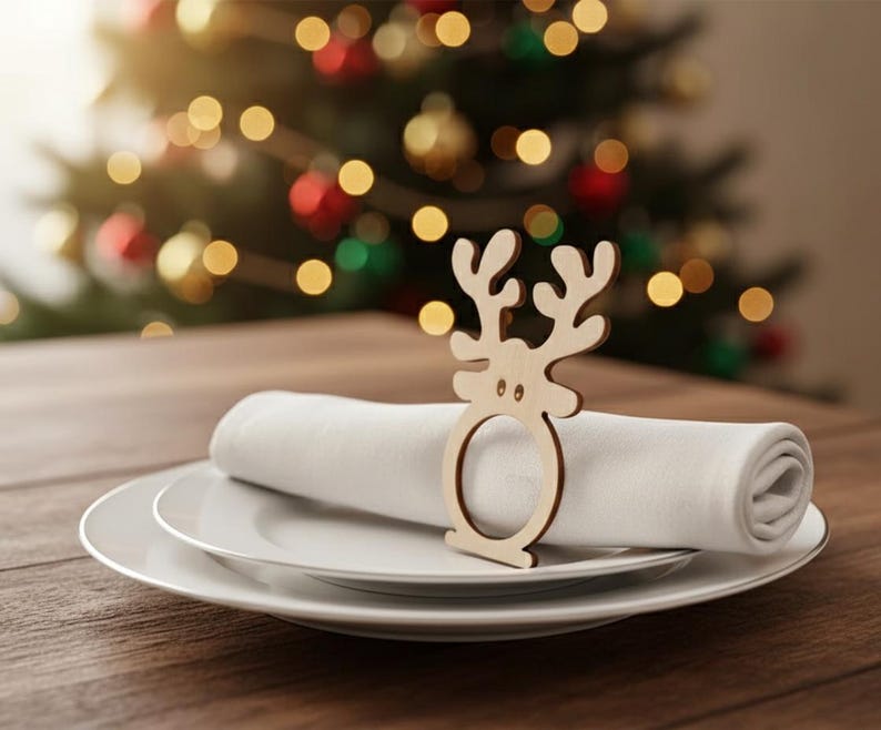 May include: A wooden reindeer napkin ring sits atop a white rolled napkin, placed on a white plate. The table setting is on a wooden surface, with a blurred Christmas tree in the background.