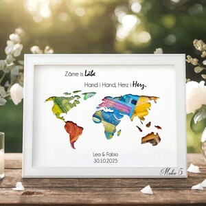 May include: A framed world map art print with colorful continents. The white frame surrounds the map, which has text at the top and bottom. The text reads "Zame is L&auml;be Hand i Hand, Herz i Herz. Lea & Fabio 30.10.2025".