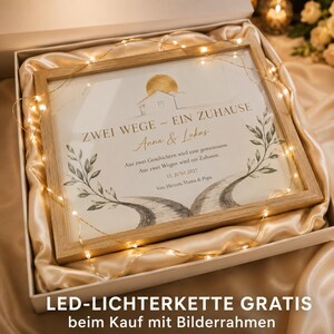 Op de afbeelding: Een lichthouten ingelijste print met een gouden zonne-illustratie, twee kronkelende paden en de Duitse tekst ZWEI WEGE - EIN ZUHAUSE, Anna & Lukas, 12. JUNI 2027, omringd door een gloeiende warmwitte LED-lichtsnoer in een geschenkdoos.