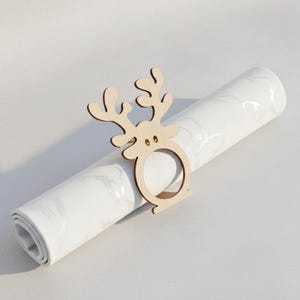 May include: A wooden reindeer napkin ring sits atop a rolled-up white napkin with a marble pattern. The reindeer has a simple design with antlers and a circular ring for the napkin. The background is a plain white surface.