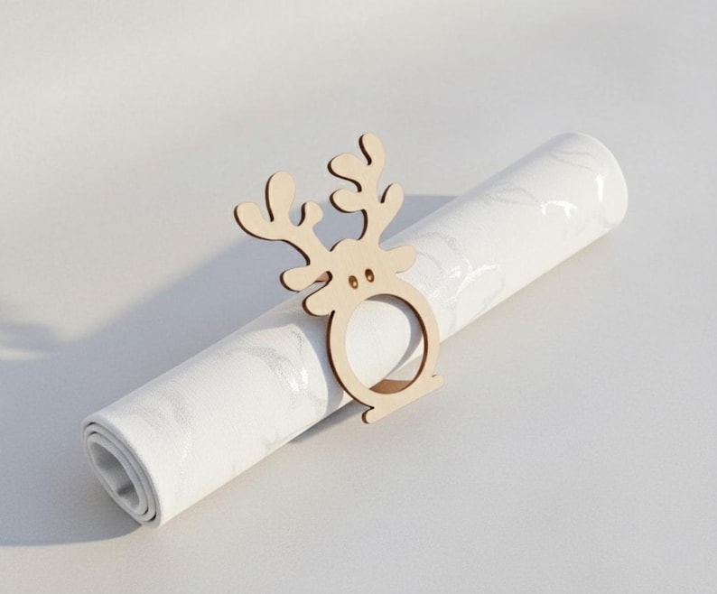 May include: A wooden reindeer napkin ring sits atop a rolled-up white napkin with a marble pattern. The reindeer has a simple design with antlers and a circular ring for the napkin. The background is a plain white surface.