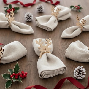 May include: A collection of beige linen napkins, each held by a wooden reindeer napkin ring. The napkins are arranged on a wooden surface, with red ribbon, holly sprigs, and pine cones scattered around, creating a festive holiday theme.