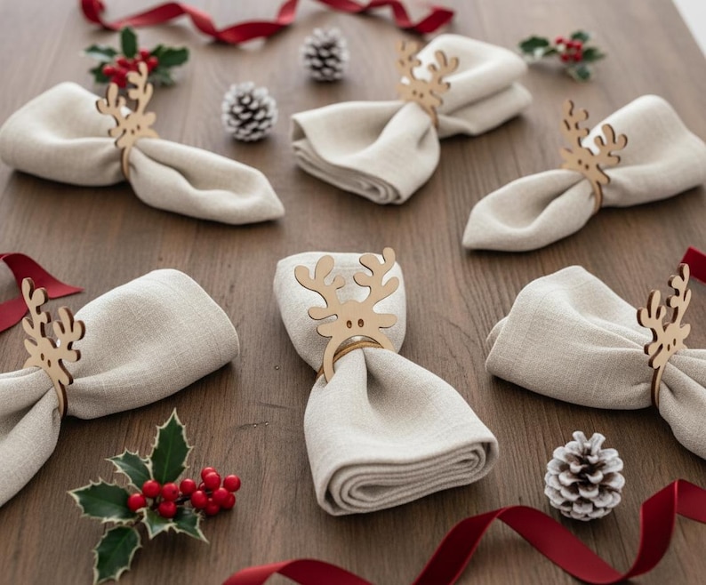 May include: A collection of beige linen napkins, each held by a wooden reindeer napkin ring. The napkins are arranged on a wooden surface, with red ribbon, holly sprigs, and pine cones scattered around, creating a festive holiday theme.