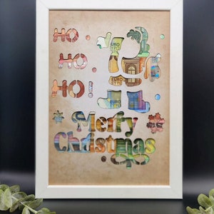 Puede incluir: Una imagen enmarcada en blanco con un fondo beige. La imagen presenta un dise&ntilde;o recortado de un reno con las palabras "&iexcl;Ho Ho Ho! &iexcl;Feliz Navidad!" en una fuente decorativa. Los recortes est&aacute;n llenos de billetes de euro.