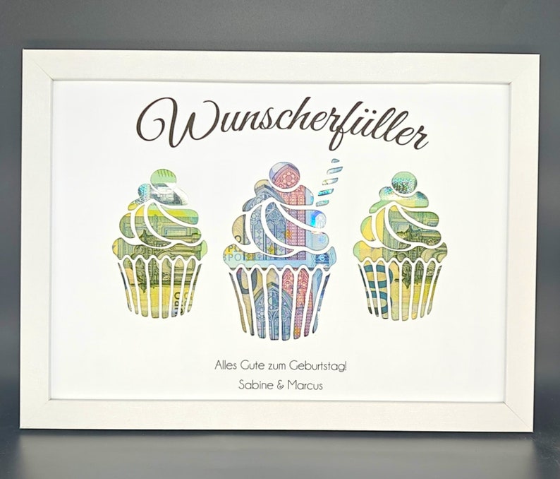 K&ouml;nnte beinhalten: Ein wei&szlig;er Bilderrahmen mit einem wei&szlig;en Passepartout und einem wei&szlig;en Hintergrund. Der Text "Wunscherf&uuml;ller" befindet sich oben im Rahmen. Drei Cupcake-Ausschnitte sind mit Geld gef&uuml;llt. Der Text "Alles Gute zum Geburtstag Sabine & Marcus" befindet sich unten im Rahmen.