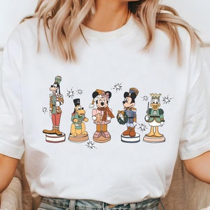 May include: White t-shirt with a graphic of Mickey Mouse, Donald Duck, Goofy, Pluto, and Minnie Mouse dressed as nutcrackers. The characters are standing on a platform with snowflakes around them.