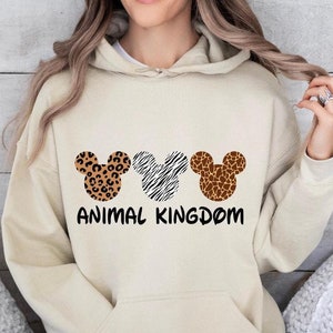 May include: A cream-colored hoodie with a graphic of three Mickey Mouse heads. Each head is patterned with a different animal print: leopard, zebra, and giraffe. The text "ANIMAL KINGDOM" is printed below the heads.