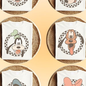 May include: Six white t-shirts with different Disney character designs. Each shirt features a checkered circle around the character's face. The characters are Mickey Mouse, Minnie Mouse, Goofy, Pluto, Donald Duck, and Daisy Duck.