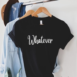 May include: Black t-shirt with the word "Whatever" in white script. The shirt is hanging on a wooden hanger. Other clothing items, including a denim shirt and a brown hat, are visible in the background.