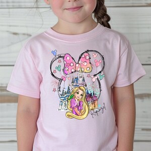Puede incluir: Camiseta rosa con un gr&aacute;fico de Rapunzel, el castillo de Cenicienta y un lazo rosa con lunares. El gr&aacute;fico est&aacute; dentro de un contorno de cabeza de Mickey Mouse.