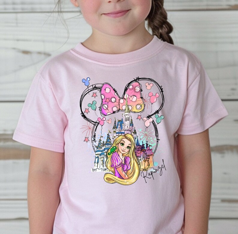 Puede incluir: Camiseta rosa con un gr&aacute;fico de Rapunzel, el castillo de Cenicienta y un lazo rosa con lunares. El gr&aacute;fico est&aacute; dentro de un contorno de cabeza de Mickey Mouse.