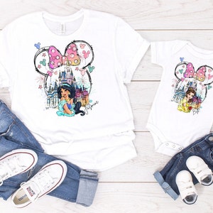 Puede incluir: Una camiseta blanca y un body blanco con una ilustraci&oacute;n colorida de una princesa Disney, un castillo y orejas de Mickey Mouse con un lazo rosa. El texto "Jasmine" est&aacute; escrito en la camiseta y "Belle" est&aacute; escrito en el body.