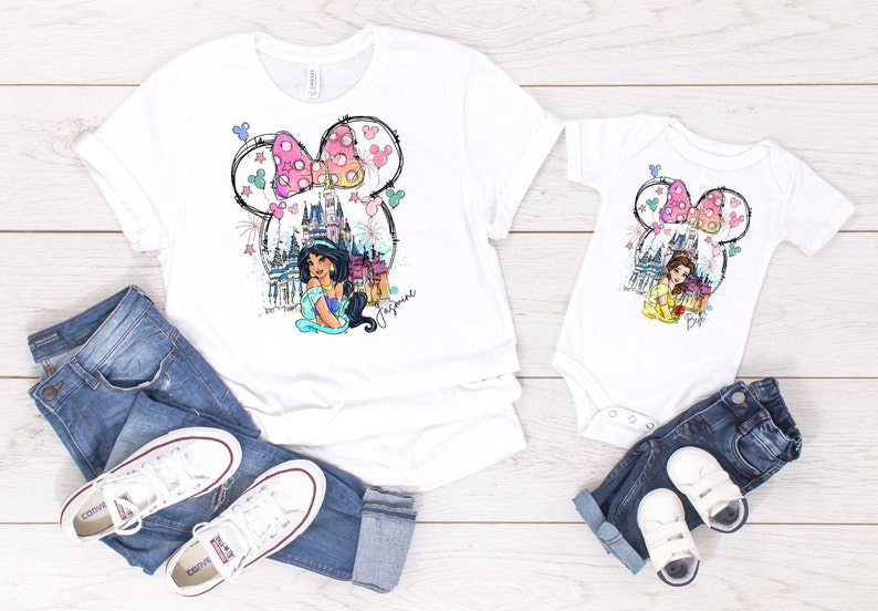 Puede incluir: Una camiseta blanca y un body blanco con una ilustraci&oacute;n colorida de una princesa Disney, un castillo y orejas de Mickey Mouse con un lazo rosa. El texto "Jasmine" est&aacute; escrito en la camiseta y "Belle" est&aacute; escrito en el body.