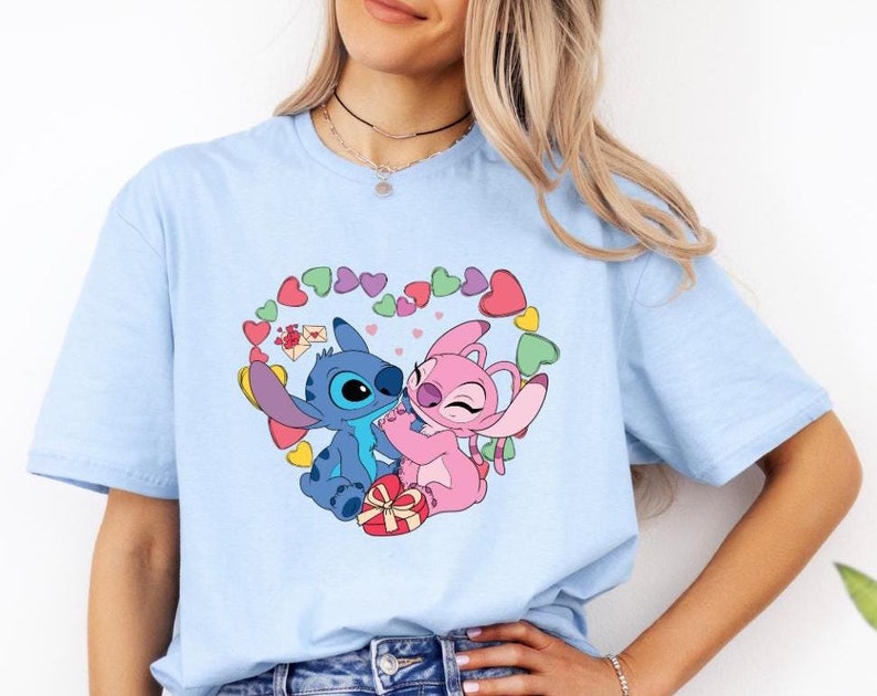 May include: Light blue t-shirt featuring a graphic of Stitch and Angel from Lilo & Stitch. The characters are in a heart shape surrounded by pink and purple hearts.