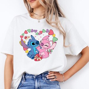 May include: White t-shirt featuring a graphic of Stitch and Angel from Lilo & Stitch. The characters are holding a gift box and surrounded by hearts.