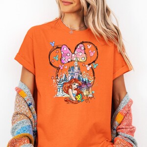 Puede incluir: Camiseta naranja con un gr&aacute;fico de un lazo rosa y blanco con lunares con orejas de Mickey Mouse, un castillo y Ariel de La Sirenita. El texto "Ariel" est&aacute; escrito debajo del gr&aacute;fico.