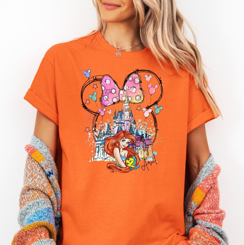 Puede incluir: Camiseta naranja con un gr&aacute;fico de un lazo rosa y blanco con lunares con orejas de Mickey Mouse, un castillo y Ariel de La Sirenita. El texto "Ariel" est&aacute; escrito debajo del gr&aacute;fico.