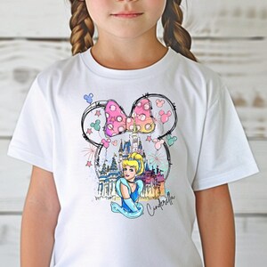 Puede incluir: Camiseta blanca con un gr&aacute;fico colorido de Cenicienta frente al Castillo de Cenicienta con orejas de Mickey Mouse y un lazo rosa. El texto "Cenicienta" est&aacute; escrito debajo del gr&aacute;fico.