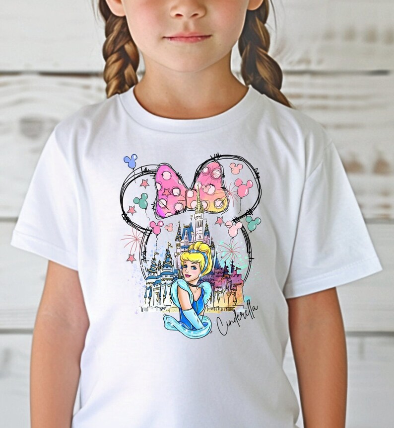 Puede incluir: Camiseta blanca con un gr&aacute;fico colorido de Cenicienta frente al Castillo de Cenicienta con orejas de Mickey Mouse y un lazo rosa. El texto "Cenicienta" est&aacute; escrito debajo del gr&aacute;fico.