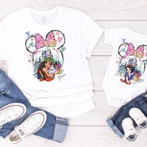 Puede incluir: Camisas blancas con una ilustraci&oacute;n colorida de una princesa de Disney, un castillo y fuegos artificiales dentro de una silueta de oreja de Mickey Mouse. El texto "Mouse" est&aacute; escrito debajo de la ilustraci&oacute;n. Las camisas se combinan con jeans azules y zapatillas blancas.