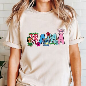 May include: A beige t-shirt with a colorful "MAMA" graphic featuring Stitch and other characters from the Disney movie Lilo & Stitch. The letters are decorated with flowers and leaves.