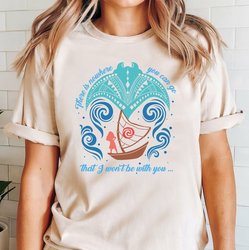Puede incluir: Una camiseta beige con un dise&ntilde;o gr&aacute;fico azul y turquesa que representa una ola estilizada con un peque&ntilde;o barco y una persona dentro. El texto "There is nowhere you can go that I won't be with you..." est&aacute; impreso debajo del gr&aacute;fico.