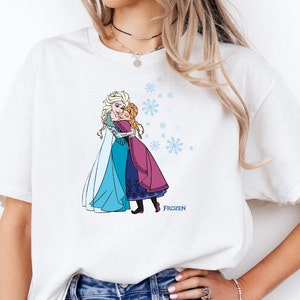 May include: White t-shirt with a graphic of the Disney Frozen characters Elsa and Anna hugging. The graphic is in color and includes snowflakes. The text "FROZEN" is printed below the graphic.