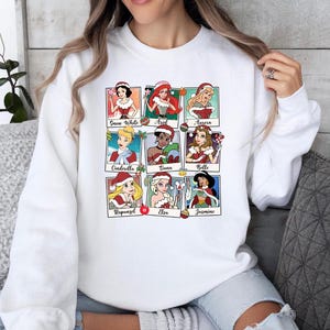 Puede incluir: Sudadera blanca con un gr&aacute;fico colorido de princesas Disney vestidas con gorros de Pap&aacute; Noel. Las princesas incluyen Blancanieves, Ariel, Aurora, Cenicienta, Tiana, Bella, Rapunzel, Elsa y Jasmine.