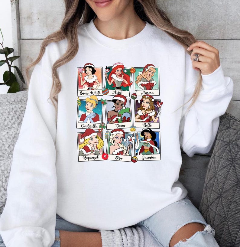 Puede incluir: Sudadera blanca con un gr&aacute;fico colorido de princesas Disney vestidas con gorros de Pap&aacute; Noel. Las princesas incluyen Blancanieves, Ariel, Aurora, Cenicienta, Tiana, Bella, Rapunzel, Elsa y Jasmine.