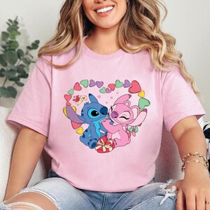 May include: Pink t-shirt featuring a graphic of Stitch and Angel from Lilo & Stitch. The graphic depicts Stitch and Angel embracing, surrounded by a heart shape made of pink and green hearts.